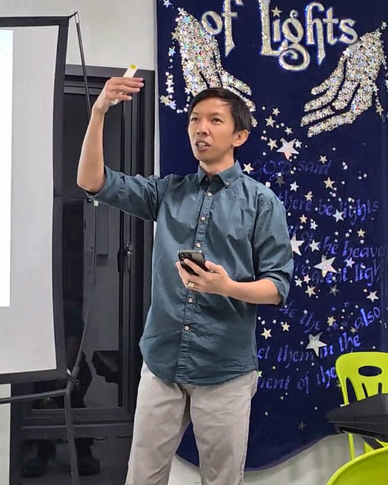 Ptr. Edwin Joseph &ldquo;Jeff&rdquo; Villagomez preaching, one hand raised toward the screen with a presentation clicker, a &ldquo;Father of Lights&rdquo; banner behind him.